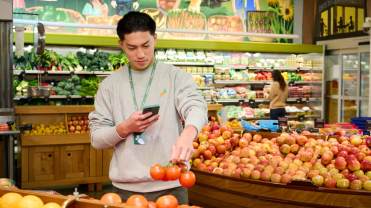 Photograph of a man picking up produce at a store and taking a picture of it with his phone.