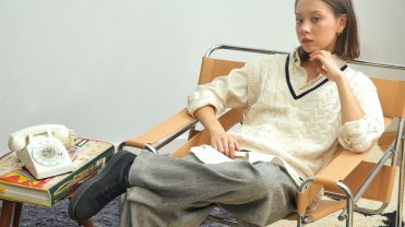 Model in a white sweater and gray pants sits in a tan lounge chair showing off black Atoms sneakers.