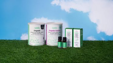 Two white containers of baby formula, and two cartons of vitamin d and probiotic drops, placed on a bed of grass with a sky-blue background