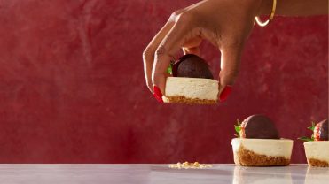 A hand selecting a strawberry-covered cheese back bite on a red background