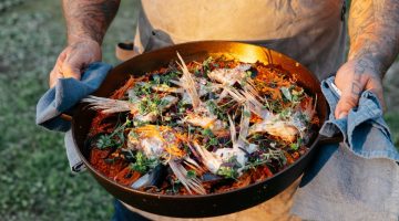 Man with tattoos on arms in tan apron holding a cast iron pan filled with food