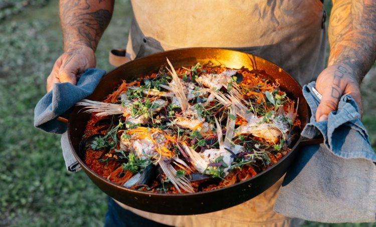 Man with tattoos on arms in tan apron holding a cast iron pan filled with food