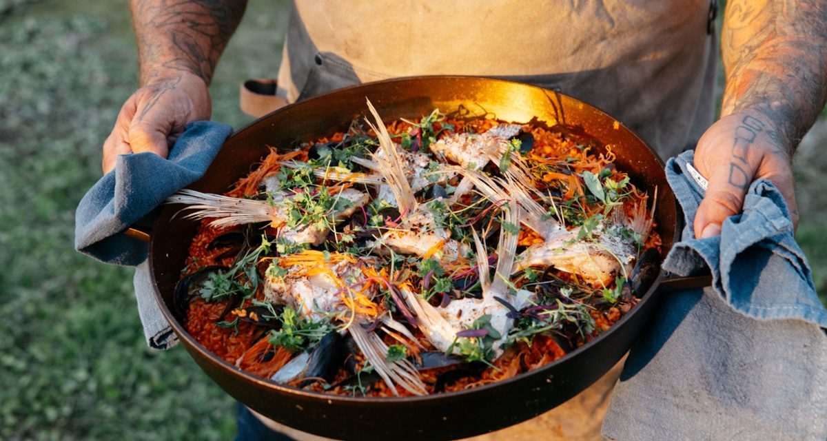 Man with tattoos on arms in tan apron holding a cast iron pan filled with food