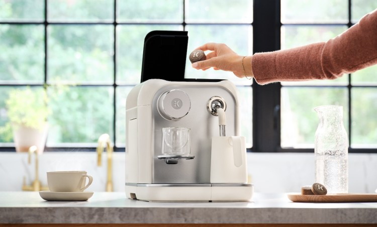 A person places a compressed coffee tablet inside a Keurig Alta machine sitting on a counter.